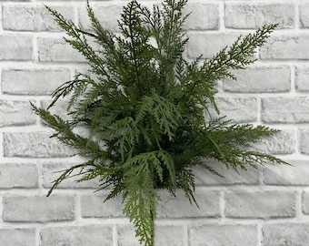 May include: A decorative artificial cedar branch featuring dense, dark green needle-like foliage arranged in a fan-like spray. The botanical piece has a central stem that branches out into multiple smaller, textured sprigs of varying lengths.