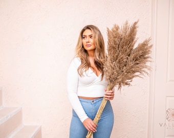 May include: A woman in a white long-sleeved top and blue jeans holds a large bouquet of dried pampas grass.
