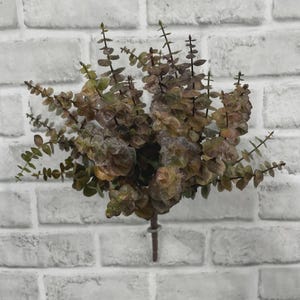 May include: An artificial eucalyptus plant stem featuring multiple branches with small, rounded, dark green and brownish-red leaves. The plant has a single central stem that extends downward, showcasing a dense, layered foliage arrangement throughout.