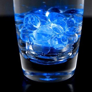 May include: A clear glass filled with water and blue gel balls. The glass is sitting on a black surface.