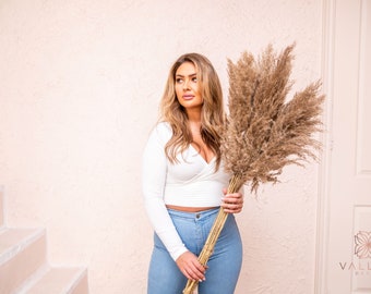 May include: A woman in a white long-sleeved top and blue jeans holds a large bouquet of dried pampas grass.