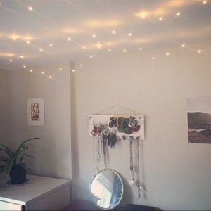 Op de afbeelding: Een witte muur met een witte houten sieradenrek met verschillende kettingen, armbanden en oorbellen die eraan hangen. Een kleine ronde spiegel met een gouden lijst hangt aan de muur onder het rek. Een kleine plant in een pot staat op een witte tafel in de hoek van de kamer. String lights zijn over het plafond gehangen.