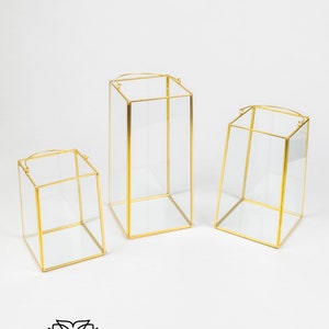 May include: Three gold-toned metal framed glass candle holders of varying sizes. The candle holders are clear glass with a gold metal frame and a hanging loop at the top.