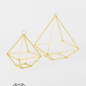May include: Two gold geometric wire frames in the shape of a pyramid. The frames are empty and are sitting on a white surface.