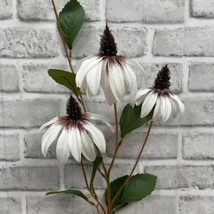 May include: A bouquet of three white artificial flowers with brown centres. The flowers are on a stem with green leaves and are arranged against a white brick wall.