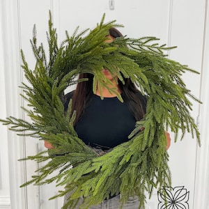 May include: A green artificial pine wreath, held in front of a person wearing a black shirt and blue jeans. The wreath is large and full, with many branches. The background is a white door.