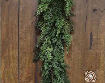 May include: A long, green garland of artificial cedar branches hangs against a rustic wooden background. The garland is perfect for adding a touch of greenery to your home decor.
