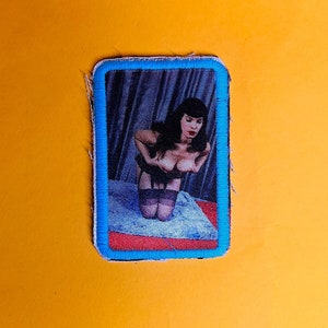 Può includere: Una toppa con cornice blu con una foto di una donna in un vestito nero e calze. La donna sta posando con le mani sui fianchi.