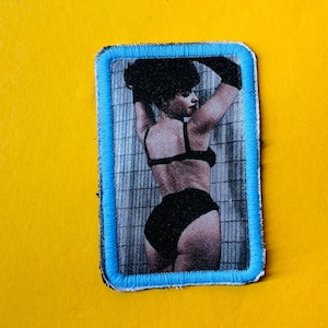 May include: A black and white photo of a woman in a black bikini, standing in a shower stall. The photo is printed on a fabric patch with a blue border.