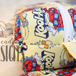 May include: A yellow fabric pouch featuring a repeating pattern of the Kool-Aid Man character in red, blue, and white. The pouch has a white zipper closure with a blue ribbon pull tab. The text Kool-Aid is printed repeatedly on the fabric.