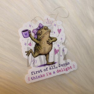 May include: A die-cut sticker featuring a tan toad wearing a purple bow, holding a purple mug, and standing near a spool of thread. The sticker includes small purple hearts and the text: first of all, Jesus thinks i'm a delight.