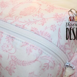 May include: Close-up of a white and pink floral patterned fabric with a white zipper. The fabric has a delicate, vintage-inspired design. The text "threadpool DESIGNS" is visible in the upper right corner.