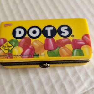 May include: A yellow rectangular box with the word "DOTS" in large, white letters. The box is filled with colorful candies and the words "A Fat Free Candy" are visible. The box has a black border and a clasp.