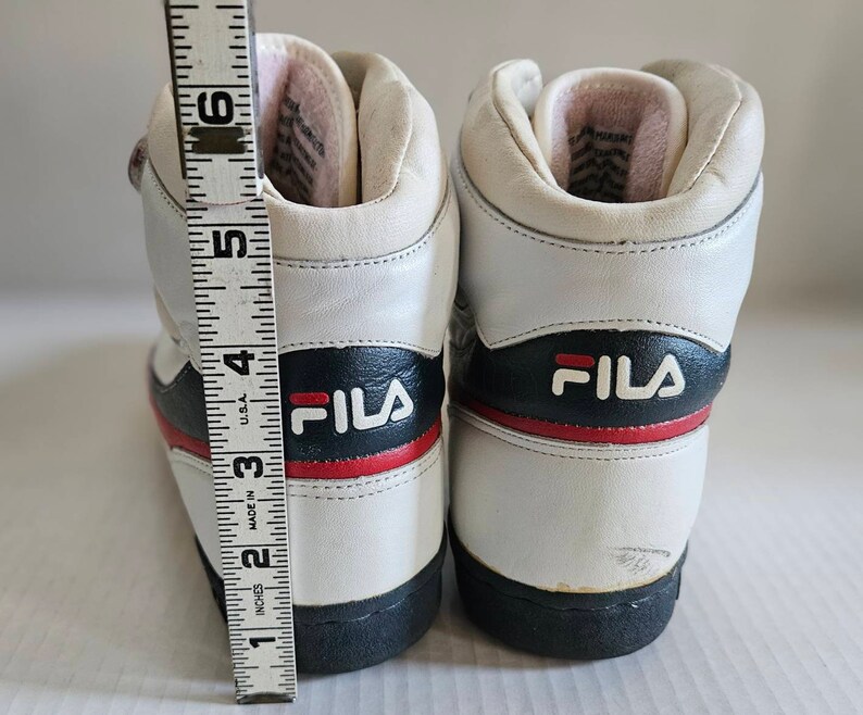 May include: A pair of white leather high-top trainers with black and red accents. The trainers have the word "FILA" printed on the side in black letters.