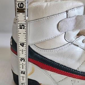 May include: White leather high-top trainers with blue and red stripes and a velcro strap. The shoes are measured with a ruler showing they are 4 inches tall.