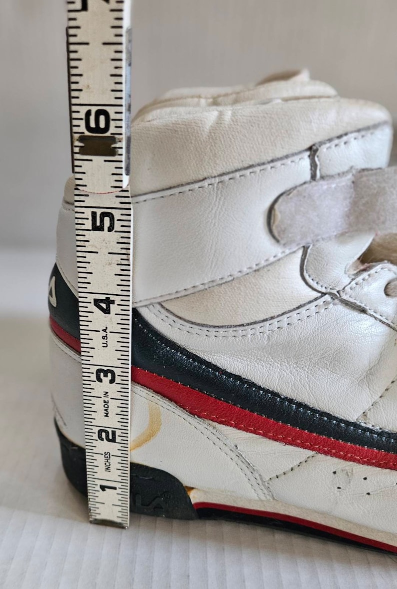 May include: White leather high-top trainers with blue and red stripes and a velcro strap. The shoes are measured with a ruler showing they are 4 inches tall.