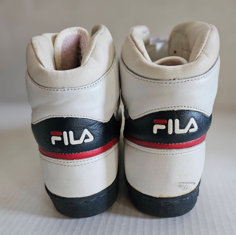 May include: A pair of white leather high-top trainers with blue and red accents. The trainers have the word "FILA" written in black on the side.