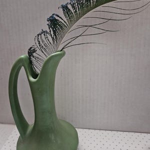 May include: A matte sage green ceramic pitcher vase with a curved handle and a flared rim. A single iridescent peacock feather with blue and green hues is placed inside the vase, extending outward from the opening of the vessel.