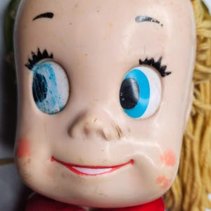 May include: A vintage doll with blonde yarn hair, a red shirt, and a smiling face with blue eyes. One eye is cracked and the other is intact.