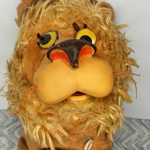 May include: A vintage plush lion toy with a brown and yellow face, large yellow eyes, and a brown nose. The lion has a long, shaggy mane and is wearing a white pull string.