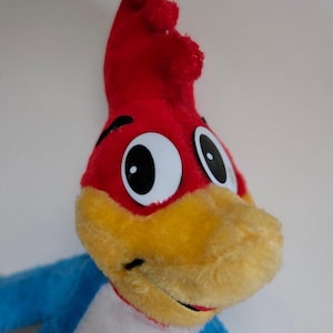 May include: A plush toy of Woody Woodpecker, a cartoon character. The toy is red, yellow, and blue. The toy has a big, round head with large, black eyes. The toy is smiling.