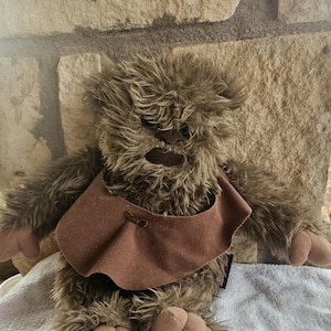 May include: A plush brown and tan furry character with a brown bib. The toy has large feet and hands, and a detailed face. The character is sitting on a white surface in front of a stone wall.