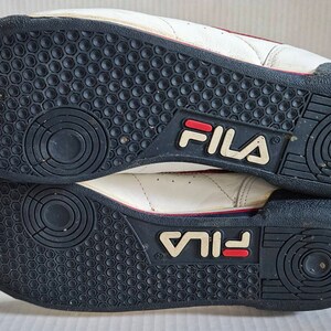 May include: A pair of white leather trainers with red and blue accents. The soles are black with a circular pattern and the word "FILA" is printed on the back of each shoe.