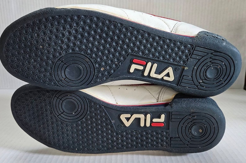 May include: A pair of white leather trainers with red and blue accents. The soles are black with a circular pattern and the word "FILA" is printed on the back of each shoe.