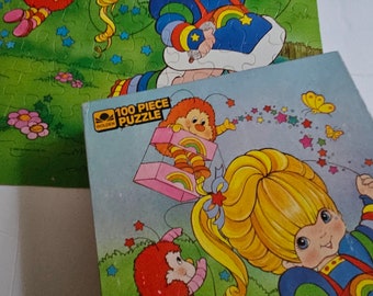Puede incluir: Un rompecabezas Golden de 100 piezas con una niña de cabello rubio y un vestido arcoíris. Está rodeada de estrellas y pequeñas criaturas rojas con alas.