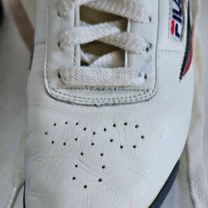 May include: White leather tennis shoes with black trim and a Fila logo on the side. The shoes have white laces and a perforated design on the toe.