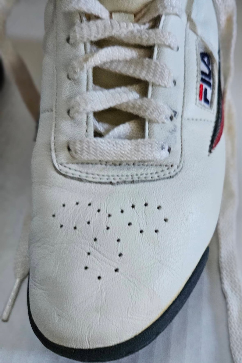 May include: White leather tennis shoes with black trim and a Fila logo on the side. The shoes have white laces and a perforated design on the toe.