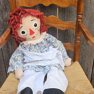 May include: A vintage rag doll with red yarn hair, a painted face, and a floral print dress, seated on a wooden chair with a woven seat. The doll wears a white apron and pants with black shoes.