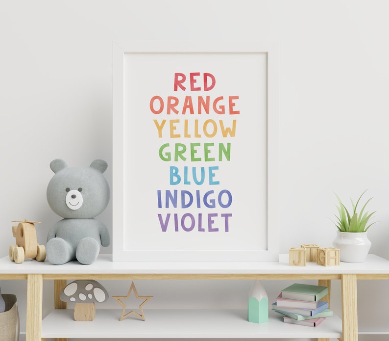 Op de afbeelding: Een wit ingelijst print met de woorden "ROOD ORANJE GEEL GROEN BLAUW INDIGO VIOLET" in regenboogkleuren. De print staat op een wit rek met een grijze teddybeer, houten speelgoed en een plant in een pot.