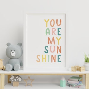 May include: A white framed print with the text "YOU ARE MY SUNSHINE" in colourful, hand-drawn letters. The print is hanging on a white wall above a wooden shelf with a grey teddy bear, a wooden mushroom, a star, a stack of books, and a plant in a white pot.