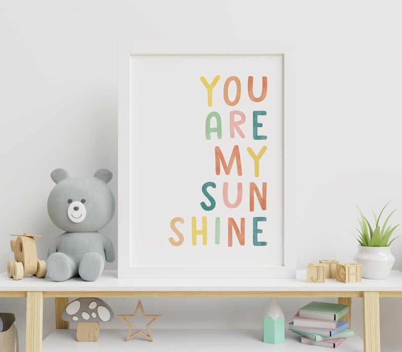 May include: A white framed print with the text "YOU ARE MY SUNSHINE" in colourful, hand-drawn letters. The print is hanging on a white wall above a wooden shelf with a grey teddy bear, a wooden mushroom, a star, a stack of books, and a plant in a white pot.