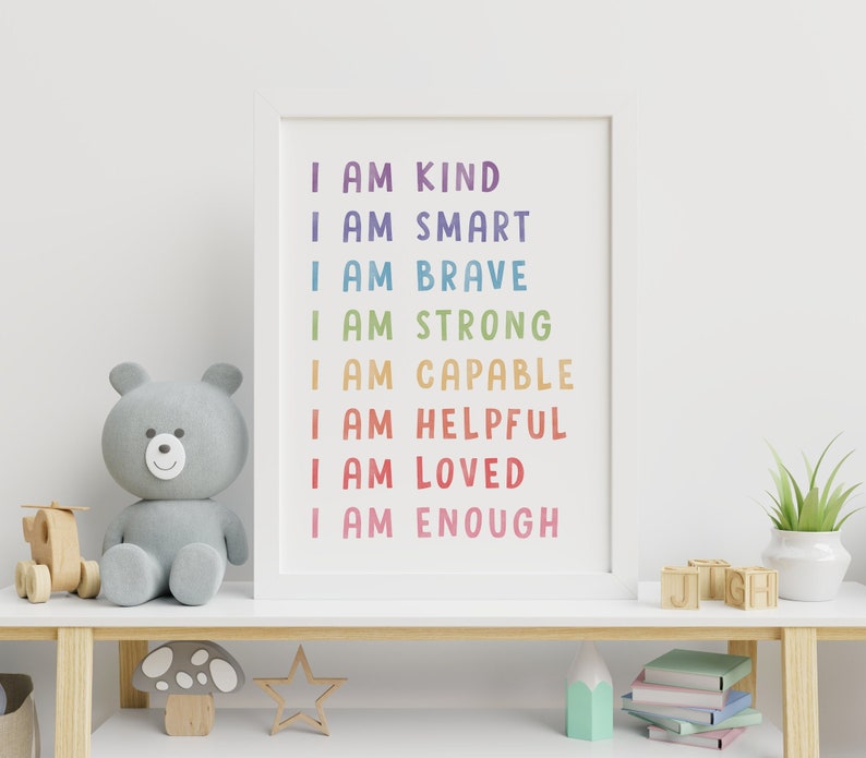 May include: A white framed print with a rainbow colored text that reads "I am kind, I am smart, I am brave, I am strong, I am capable, I am helpful, I am loved, I am enough". The print is hanging on a white wall above a wooden shelf with a gray teddy bear, a wooden toy airplane, a wooden mushroom, a star, a stack of books, and a small potted plant.