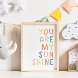 May include: A framed print with the text "You Are My Sunshine" in colourful letters. The letters are in different colours, including pink, orange, yellow, green, and blue. The print is hanging on a white wall with a garland of fabric flags.