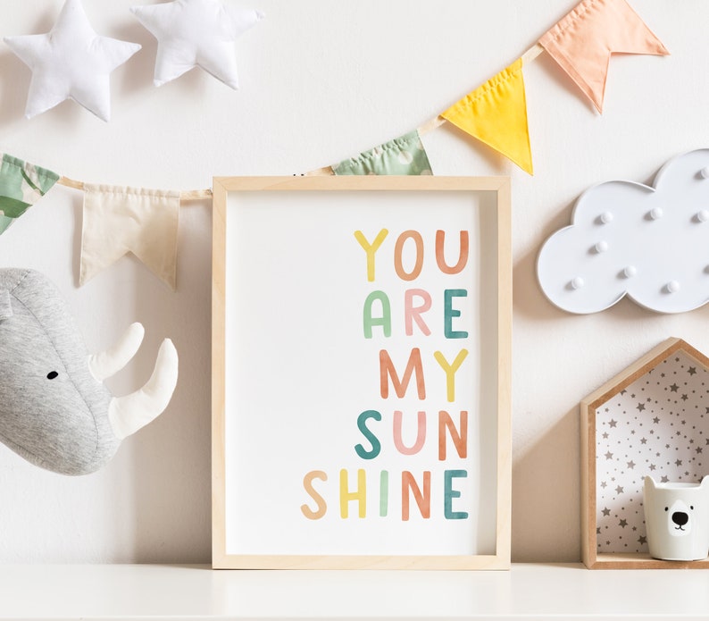 May include: A framed print with the text "You Are My Sunshine" in colourful letters. The letters are in different colours, including pink, orange, yellow, green, and blue. The print is hanging on a white wall with a garland of fabric flags.