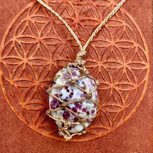 May include: A pink and white gemstone wrapped in twine, hanging from a brown cord. The gemstone is tied with a macrame knot and is resting on a brown surface with a geometric pattern.