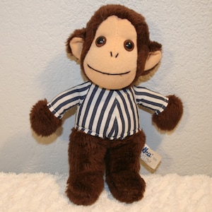 May include: A brown plush monkey wearing a blue and white striped shirt. The monkey is smiling and has brown fur. The monkey is standing on a white fluffy surface. The tag on the monkey's foot reads "Nelco".