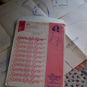 May include: A pink and white sewing pattern envelope with the text "Stretch & Sew" repeated multiple times. The envelope features a drawing of a woman wearing a sleeveless top. The text "Ear Collar Cut Four" is visible on a white pattern piece.