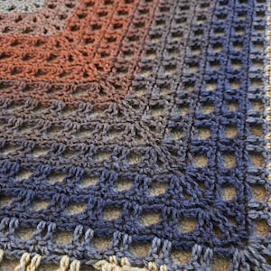 May include: Close-up of a crocheted blanket with a gradient color scheme. The blanket transitions from a warm orange-brown to a cool blue-gray, with a cream-colored border. The open weave creates a textured, airy appearance.