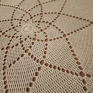 Könnte beinhalten: Nahaufnahme eines beigefarbenen, runden Häkelteppichs mit einem kreisförmigen Muster. Der Teppich hat ein zartes, spitzendes Design mit durchbrochenen Details.