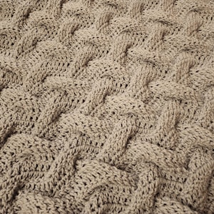 Puede incluir: Primer plano de una manta de punto beige con un patrón tejido. La manta tiene una superficie suave y texturizada.