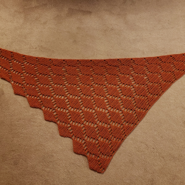 May include: A crocheted shawl in a rust orange color with a geometric pattern. The shawl is triangular in shape and has a scalloped edge.
