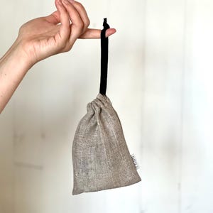 May include: A small, beige linen drawstring bag with a black drawstring. The bag is hanging from a finger.