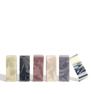 May include: Six bars of handmade soap in various colors and patterns, including green, black, pink, beige, and purple. The bars are arranged in a row, with one bar leaning forward.