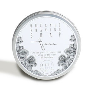 Può includere: Una scatola di latta argentata con un'etichetta bianca su cui è scritto "ORGANIC SHAVING SOAP" e "Tjåra" in corsivo. L'etichetta include anche il testo "Artisan pine tar shave soap crafted in the woods of Varmland" e un logo che dice "KALI lower Organics".