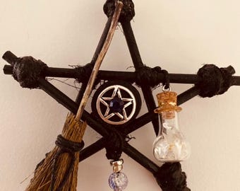 May include: A black pentagram-shaped wall hanging with a small broom, a bottle with a cork top, and a silver star pendant. The pentagram is made of dark twigs tied together with black twine. A string is attached for hanging.
