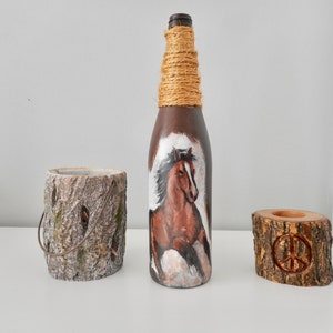 May include: A brown glass bottle with a horse painted on it, wrapped in twine. The bottle is next to two wooden candle holders, one with a peace sign carved into it.